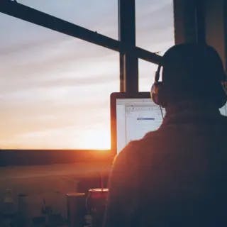 Man wearing headphones and looking at computer screen in front of sunset view through window