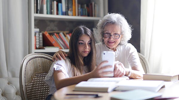 Grandmother with arm around granddaughter sitting at dining table, showing her something on a mobile phone