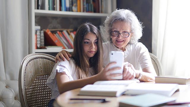 Please upGrandmother with arm around granddaughter sitting at dining table, showing her something on a mobile phonedate alt text here
