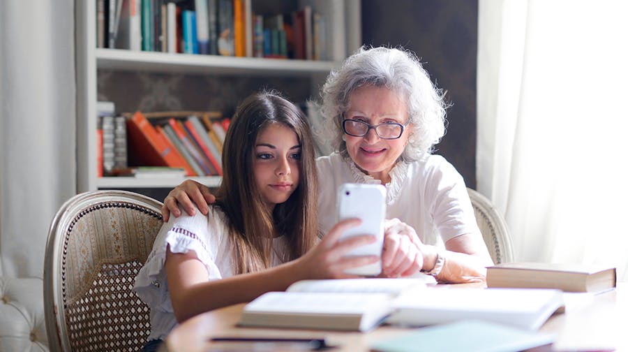 Grandmother with arm around granddaughter sitting at dining table, showing her something on a mobile phone