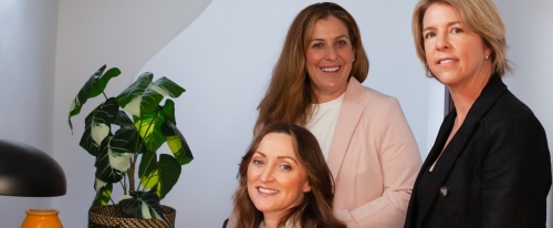 Chailee, Ainslie and Rebecca standing and sitting around a desk, smiling at camera