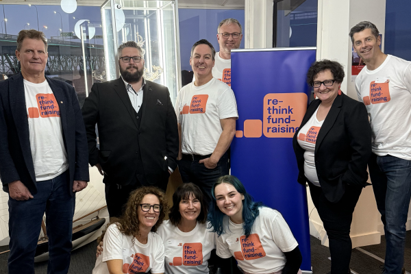 A group of people stand and sit around a branded banner smiling. They all wear white shirts that have a coral thought bubble that reads 'rethink fundraising' in dark blue lettering.